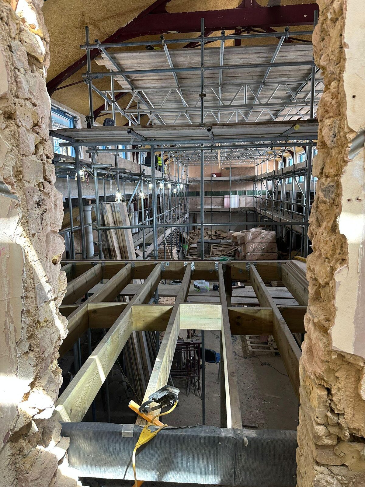 Retrofit works inside the building: scaffolding and new timber joists seen through an opening in the original wall